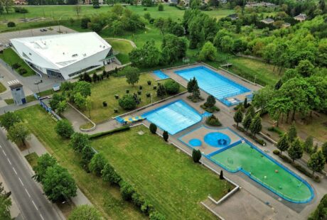 Widok z lotu ptaka na kompleks odkrytych basenów z wieloma basenami, zielonymi trawnikami, drzewami i dużym białym budynkiem basenów krytych w pobliżu.