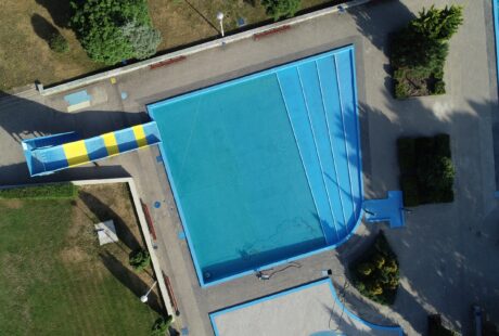 Widok z lotu ptaka na odkryty basen z żółto-niebieską zjeżdżalnią wodną, otoczony brukowanymi chodnikami i zielenią.