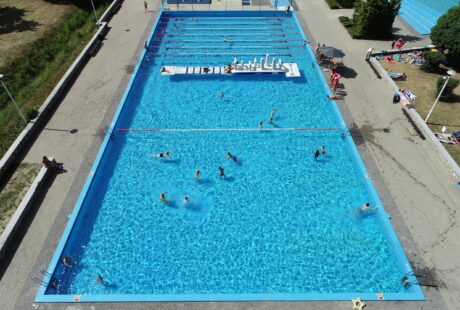 Widok z lotu ptaka na odkryty basen z ludźmi pływającymi i relaksującymi się; widoczne są tory basenowe i pływający tor przeszkód.