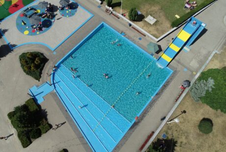 Widok z lotu ptaka na odkryty basen z pływającymi ludźmi, żółto-niebieską zjeżdżalnią, pobliskimi ławkami, zielenią i kolorowym placem zabaw.