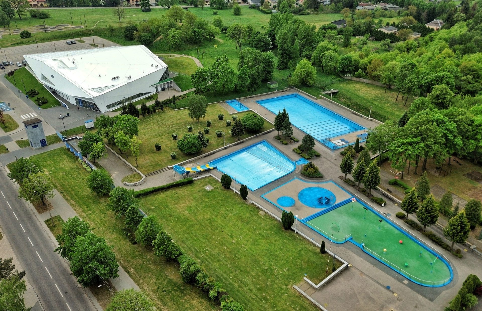 Widok z lotu ptaka na kompleks odkrytych basenów z wieloma basenami, zielonymi trawnikami, drzewami i dużym białym budynkiem basenów krytych w pobliżu.