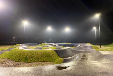 Pumptrack z gładkimi rampami i trawiastymi terenami jest oświetlony nocą przez wysokie latarnie uliczne.