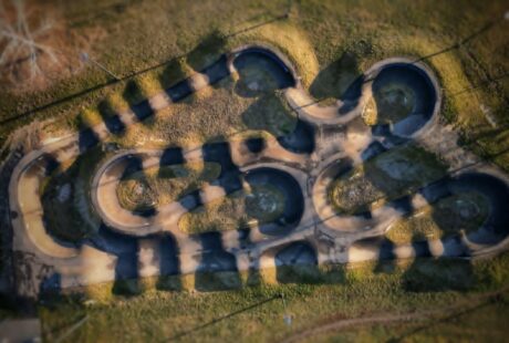 Widok z lotu ptaka na utwardzony tor rowerowy typu pumptrack z wieloma pętlami i zakrętami, otoczony trawą i rzucający długie cienie.
