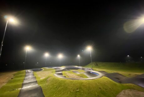 Dobrze oświetlony tor typu pumptrack na świeżym powietrzu z krętymi asfaltowymi ścieżkami i trawiastymi kopcami jest widoczny w nocy pod wieloma latarniami ulicznymi.