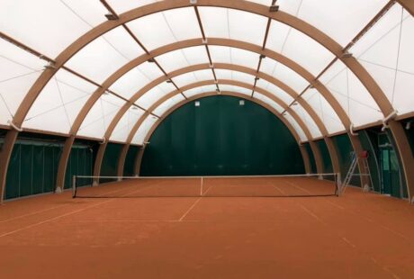 Kryty kort tenisowy z wysokim łukowatym sufitem, zielonymi ścianami i siatką rozciągniętą na środku.
