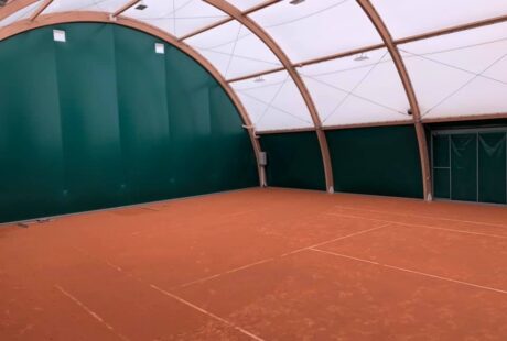 Kryty kort tenisowy z ceglaną nawierzchnią, wysokimi łukowatymi sufitami, zielonymi ścianami i górnym oświetleniem. Kort jest pusty i dobrze oświetlony.