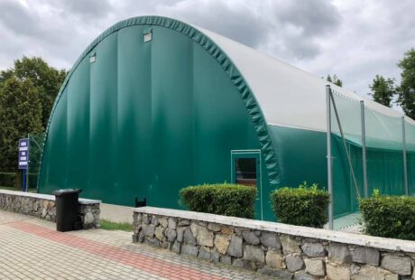 Duża zielona konstrukcja zadaszenia kortów tenisowych stoi za niskim kamiennym murem z krzewami, obok brukowanego chodnika pod zachmurzonym niebem.