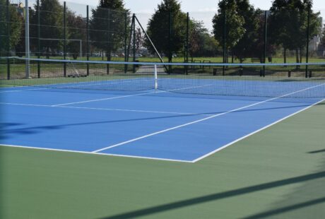 Pusty odkryty kort tenisowy z niebieską nawierzchnią i zielonym obramowaniem, otoczony drucianym ogrodzeniem i drzewami w tle.