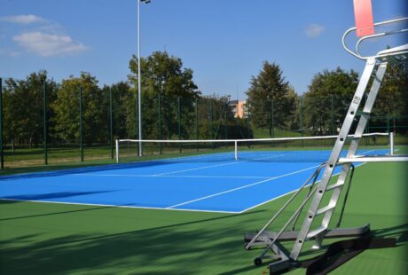 Niebiesko-zielony odkryty kort tenisowy z metalowym krzesłem sędziowskim na pierwszym planie, otoczony płotem i drzewami pod gołym niebem.