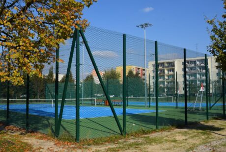 Odkryty kort tenisowy z niebiesko-zieloną nawierzchnią, otoczony wysokim zielonym płotem, z budynkami mieszkalnymi i drzewami widocznymi w tle.