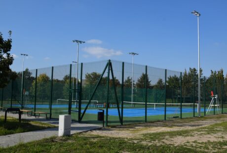 Odkryte korty tenisowe otoczone wysokim zielonym płotem, z ławkami, słupami oświetleniowymi, krzesłem sędziowskim i drzewami widocznymi w tle pod błękitnym niebem.