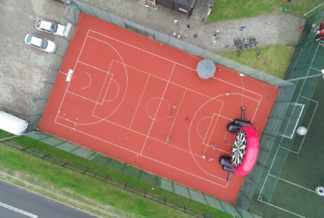 Widok z lotu ptaka na czerwone boisko wielofunkcyjne, na którym grają ludzie, otoczone ogrodzeniem; po jednej stronie znajduje się nadmuchiwana konstrukcja i parasol, a w pobliżu zaparkowane są samochody.
