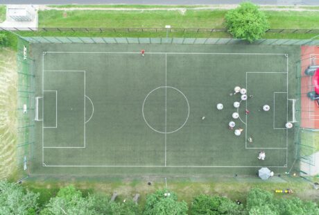 Widok z lotu ptaka na boisko do piłki nożnej z grupą ludzi zgromadzonych w pobliżu jednej bramki i pojedynczą osobą stojącą samotnie w pobliżu drugiego końca.