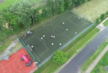 Widok z lotu ptaka na ogrodzone boisko do piłki nożnej z ludźmi w nadmuchiwanych bańkach, obok boiska do koszykówki i boiska trawiastego.