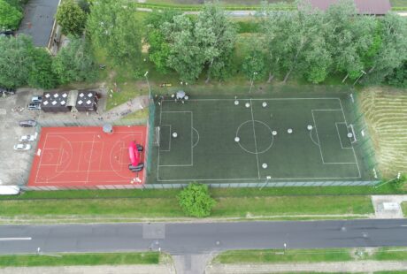 Widok z lotu ptaka na sąsiadujące odkryte boiska sportowe: czerwone boisko do koszykówki po lewej i zielone boisko do piłki nożnej po prawej, otoczone drzewami i zaparkowanymi w pobliżu pojazdami.