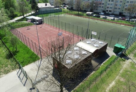 Ogrodzony zewnętrzny obiekt sportowy z czerwonym boiskiem do koszykówki, zielonym boiskiem do piłki nożnej, małym budynkiem oraz otaczającymi drzewami i budynkami mieszkalnymi.