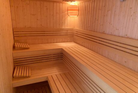 Wnętrze drewnianej sauny z wielopoziomowymi ławkami, drewnianymi zagłówkami, lampą na ścianie i termometrem w prawym górnym rogu.