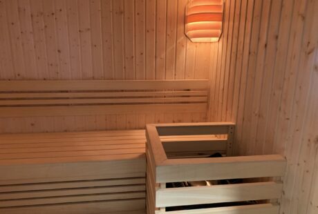 Drewniane wnętrze sauny z ławkami i oświetleniem na ścianie. Piec jest zamknięty w drewnianej ramie w rogu.