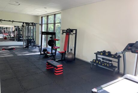Dobrze oświetlona siłownia z maszynami siłowymi, bieżnią, wolnymi ciężarami, ławkami, piłką stabilizacyjną i dużym oknem wpuszczającym naturalne światło.
