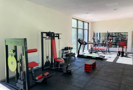 Siłownia z różnorodnym sprzętem do ćwiczeń, w tym maszynami siłowymi, wolnymi ciężarami, bieżnią, wioślarzem i platformami do aerobiku, z naturalnym światłem z dużych okien.