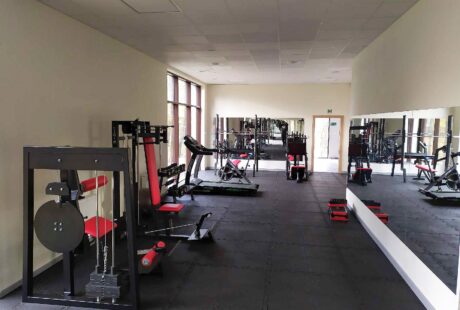Siłownia z różnymi urządzeniami do ćwiczeń, czerwonymi i czarnymi ławkami, lustrzaną ścianą, gumową podłogą i dużymi oknami zapewniającymi naturalne światło.