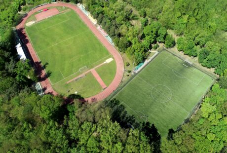 Widok z lotu ptaka na dwa sąsiadujące boiska do piłki nożnej, jedno z bieżnią i drugie otoczone drzewami, wykonane w świetle dziennym.