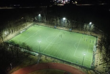Widok z lotu ptaka na oświetlone boisko do piłki nożnej ze sztuczną trawą w nocy, z graczami na boisku i bieżnią otaczającą część boiska.