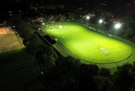 Widok z lotu ptaka na jasno oświetlone boisko do piłki nożnej w nocy, z kortem tenisowym po lewej stronie i otaczającymi drzewami i budynkami.